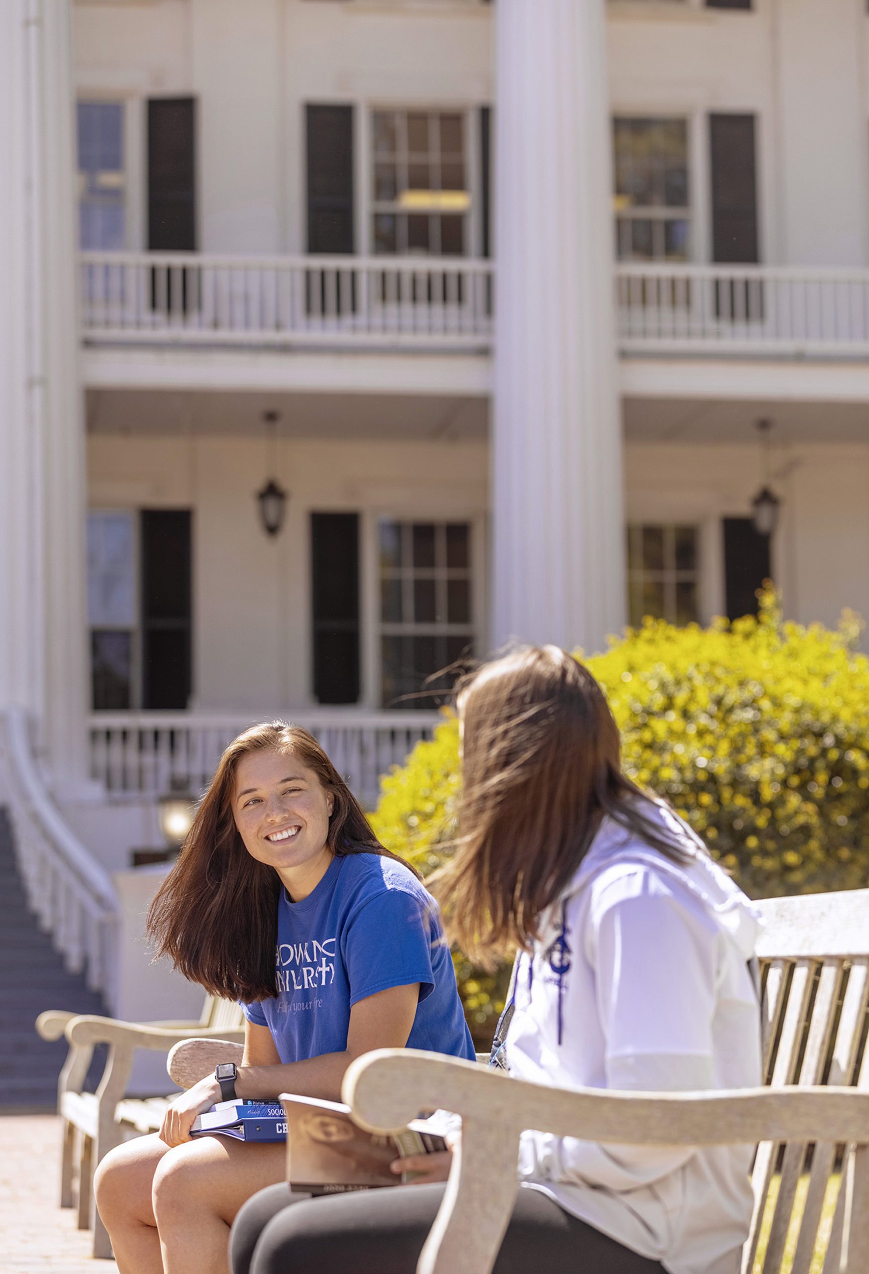 Two female students sit on a bench in front of the Columns Building